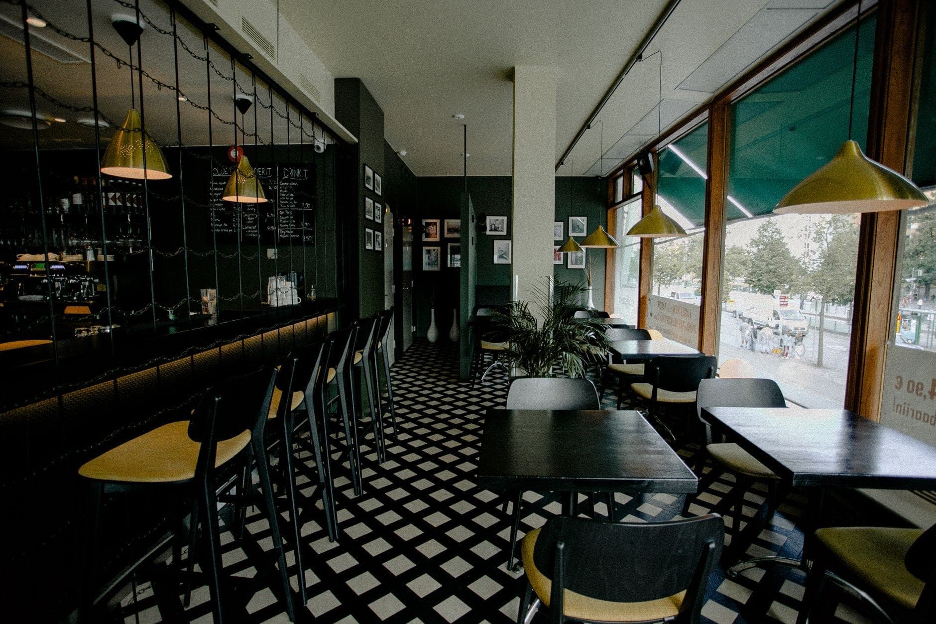 Modernes Bistro-Interieur in Warschau mit großen Fenstern, schwarz-weißem Fliesenboden, schwarzen Tischen und Stühlen sowie goldenen Pendelleuchten.