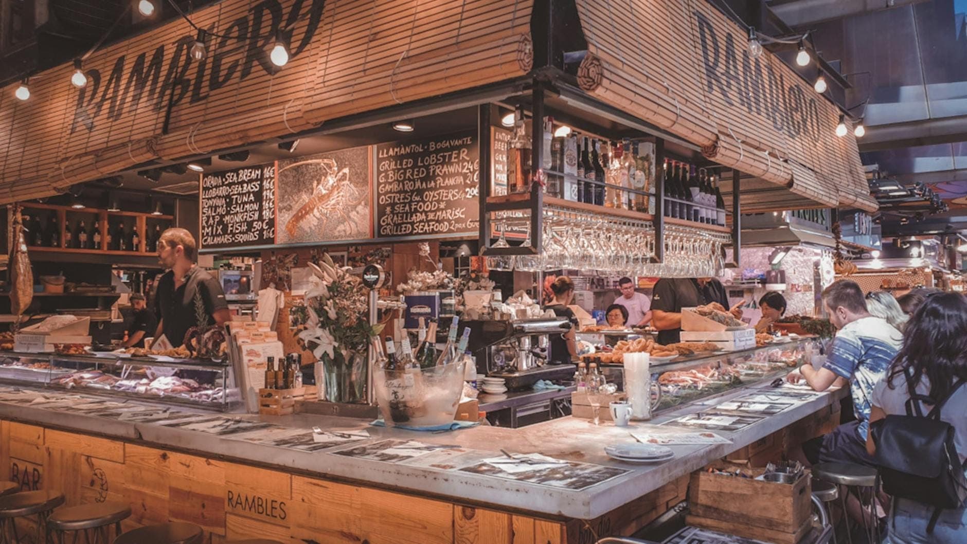 Quầy ẩm thực sôi động tại chợ Barcelona với thực khách đang dùng bữa, thực đơn viết tay, ly treo lơ lửng và không khí địa phương rộn ràng — khắc họa đúng tinh thần ẩm thực đặc sắc của thành phố.