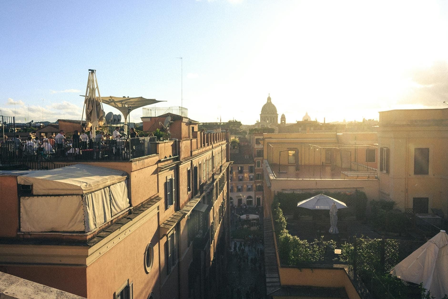 Nhà hàng trên sân thượng ở Rome lúc hoàng hôn, thực khách đang thưởng thức bữa ăn với nền là những tòa nhà cổ kính và mái vòm dưới bầu trời rực sáng.