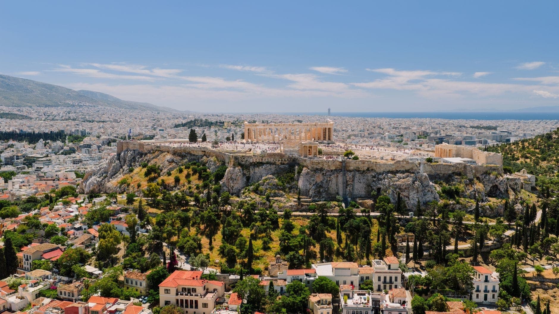 Vista aérea de la Acrópolis y los barrios circundantes, incluyendo zonas verdes, edificios residenciales y el paisaje urbano bajo un cielo azul.