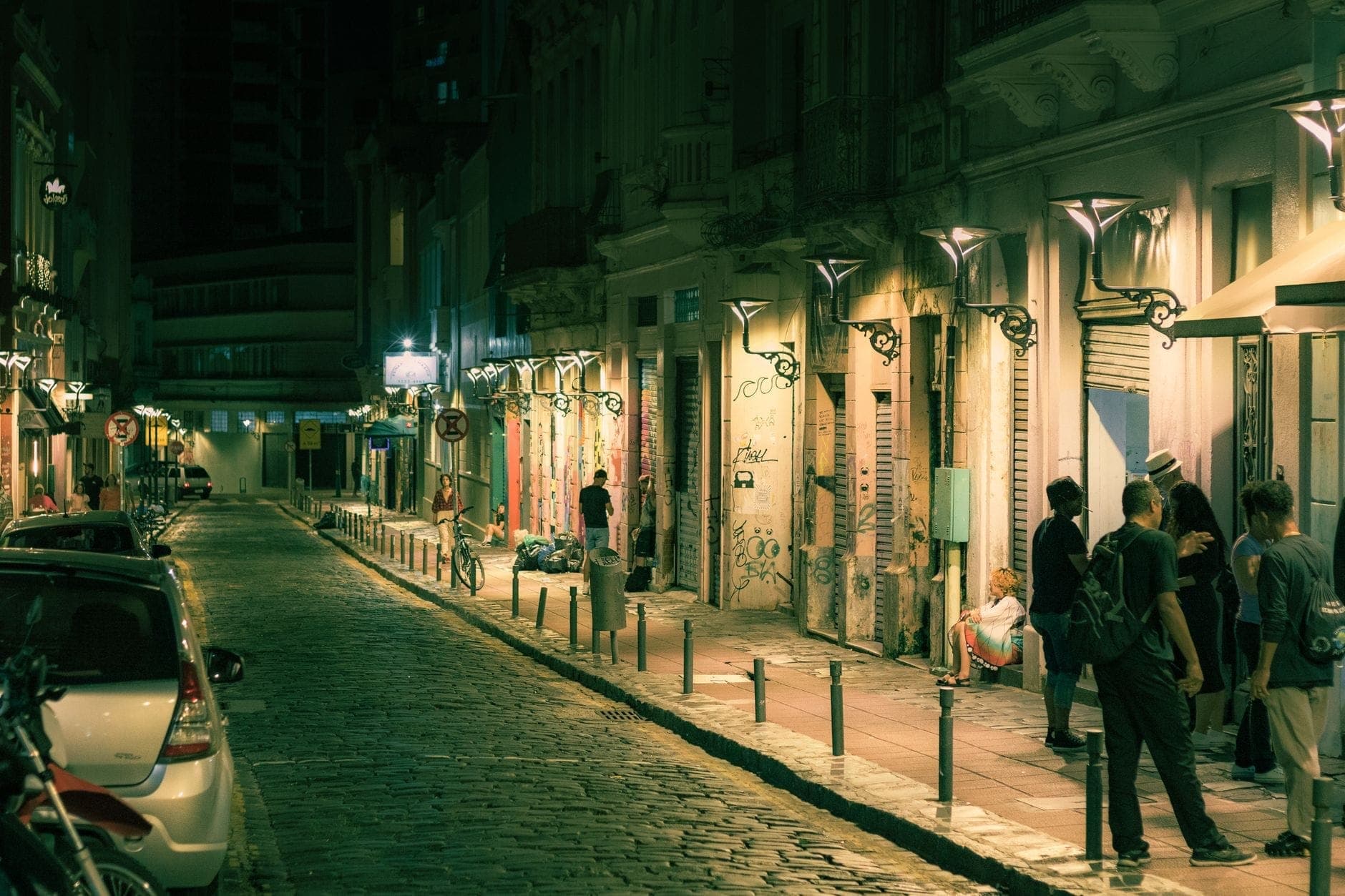 Escena nocturna de una animada calle de Atenas bordeada de tiendas iluminadas, grafitis y grupos de personas socializando en la acera.