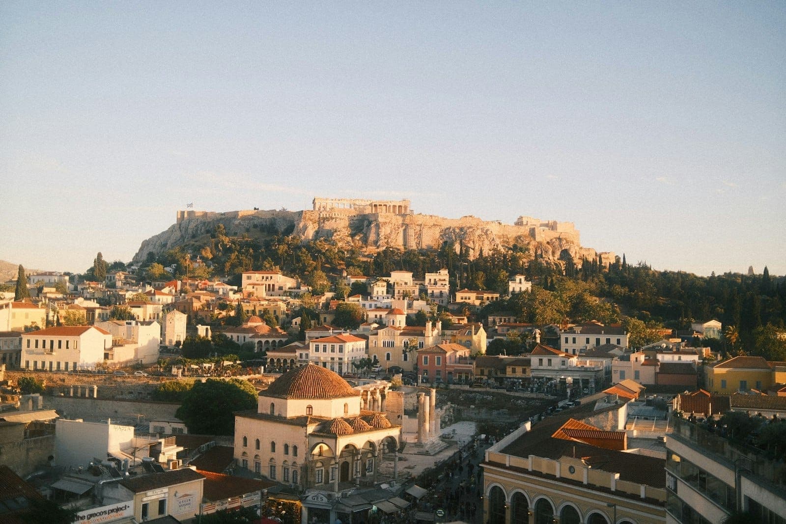 Wide view of central Athens with the Acropolis rising above city rooftops at golden hour, capturing urban neighborhoods and iconic landmarks under a clear sky.