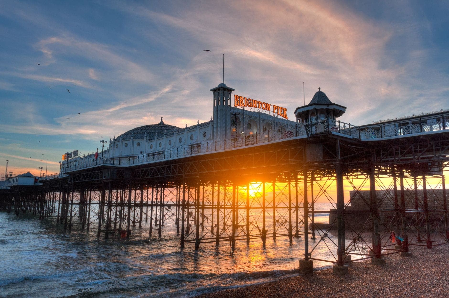 Cầu Brighton Pier lúc hoàng hôn, nắng vàng chiếu xuyên qua cầu, sóng nhẹ vỗ bờ và bầu trời rực rỡ sắc màu phía trên.