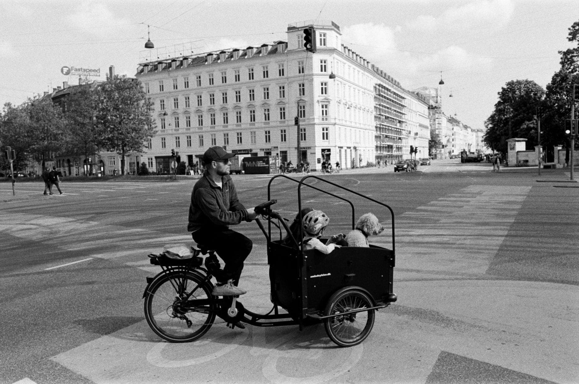 Schwarz-Weiß-Foto einer Person, die mit zwei Kindern in einem Lastenrad eine städtische Kreuzung mit historischen Gebäuden überquert.