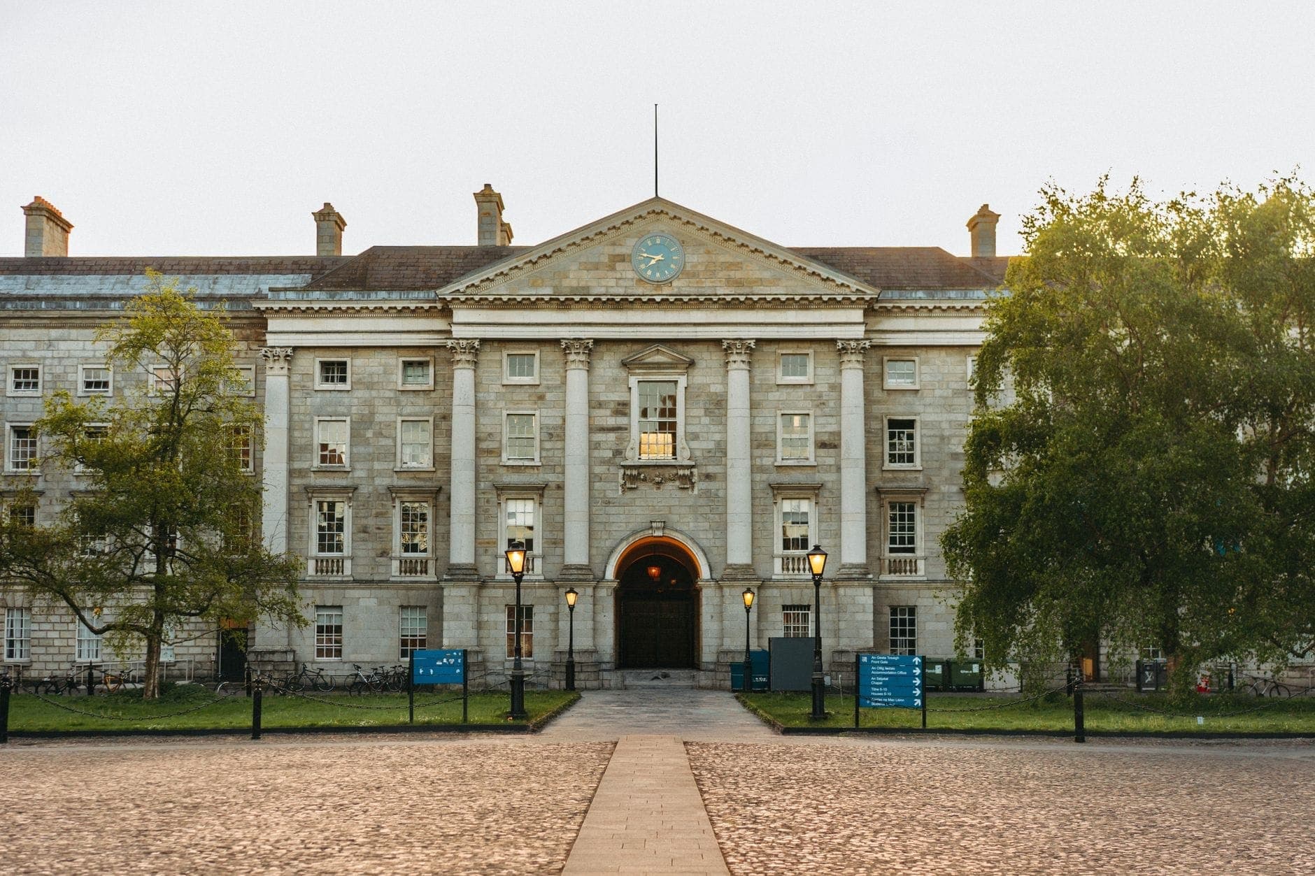 Vista frontal de la histórica fachada de piedra del Trinity College Dublin con farolas y árboles en primer plano.
