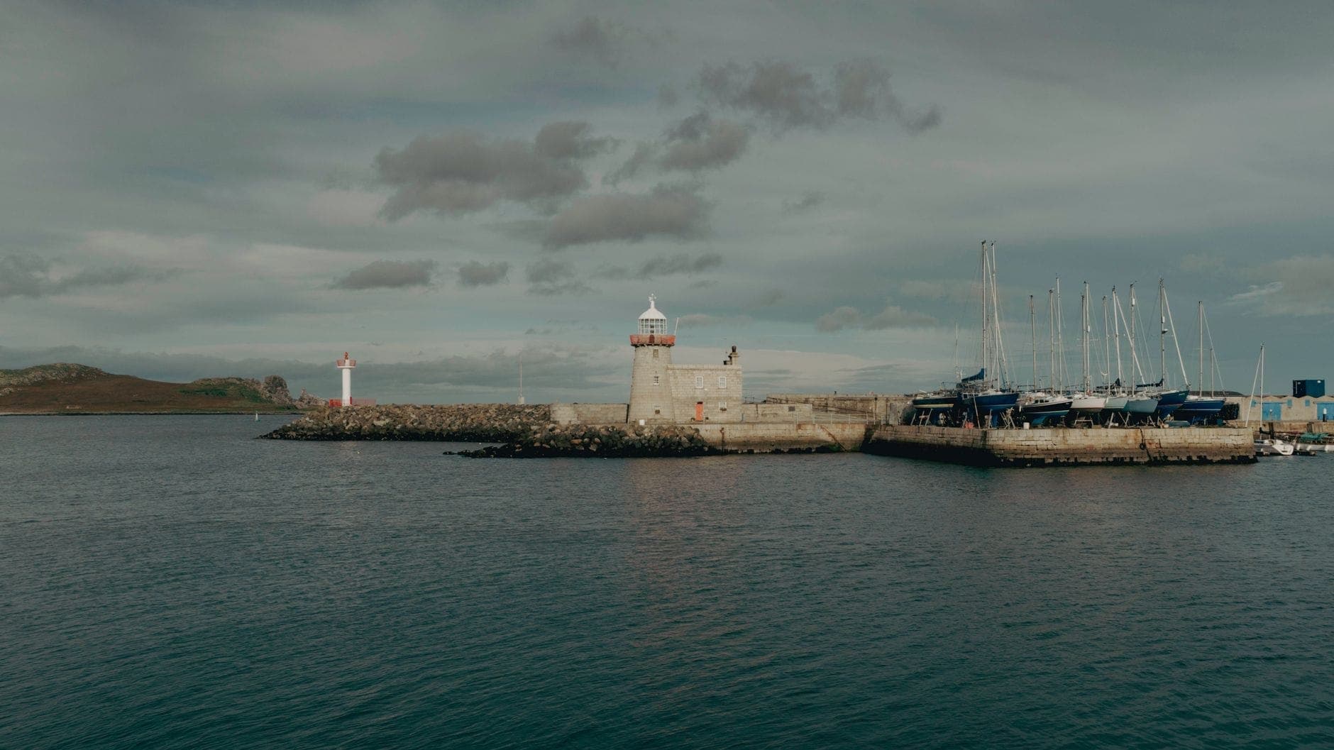Vista amplia del puerto de Howth con su emblemático faro, muelles de piedra y veleros amarrados en un día nublado.