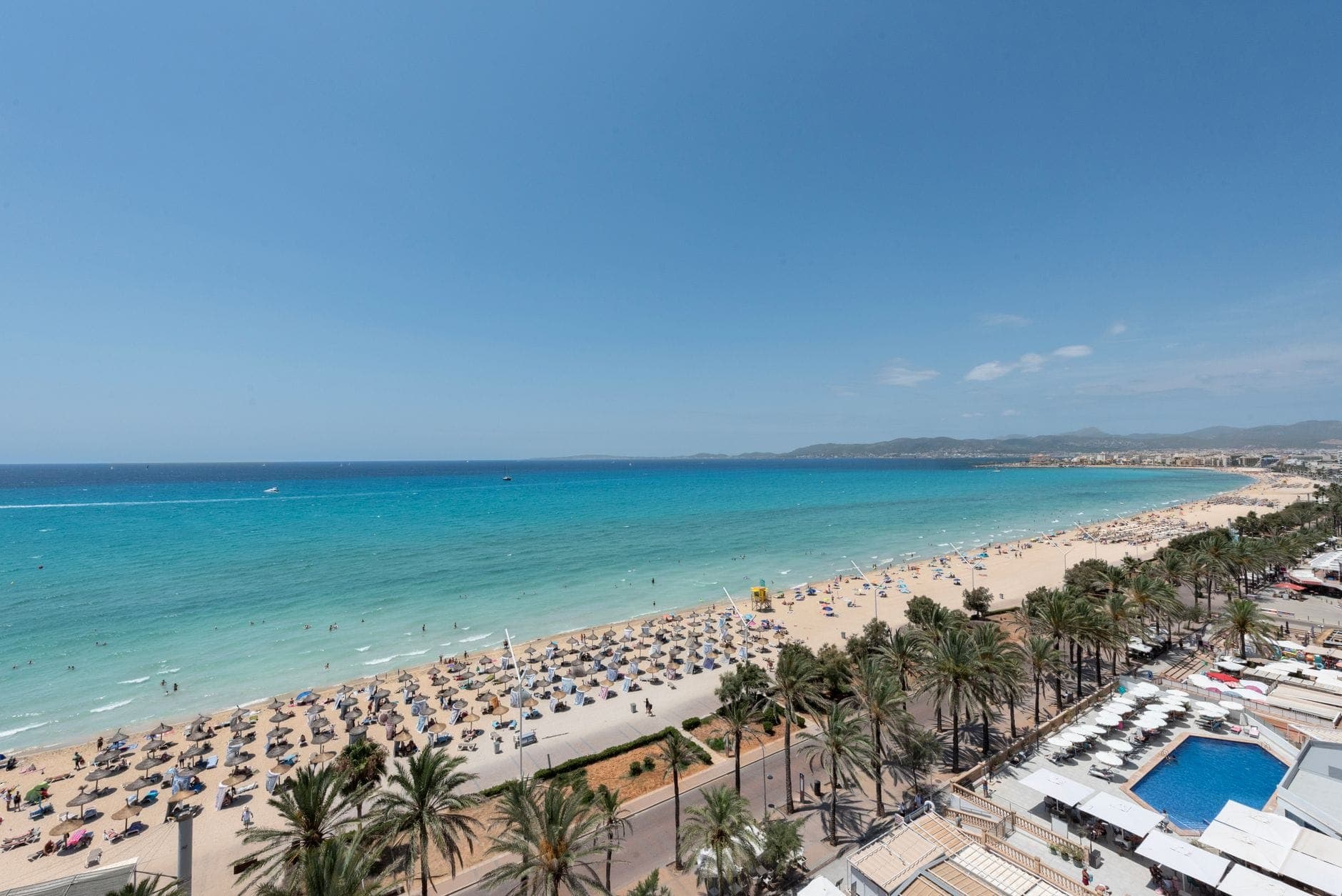 Aerial view of a long sandy beach lined with palm trees, sunbeds, and clear turquoise water next to beachfront hotels.