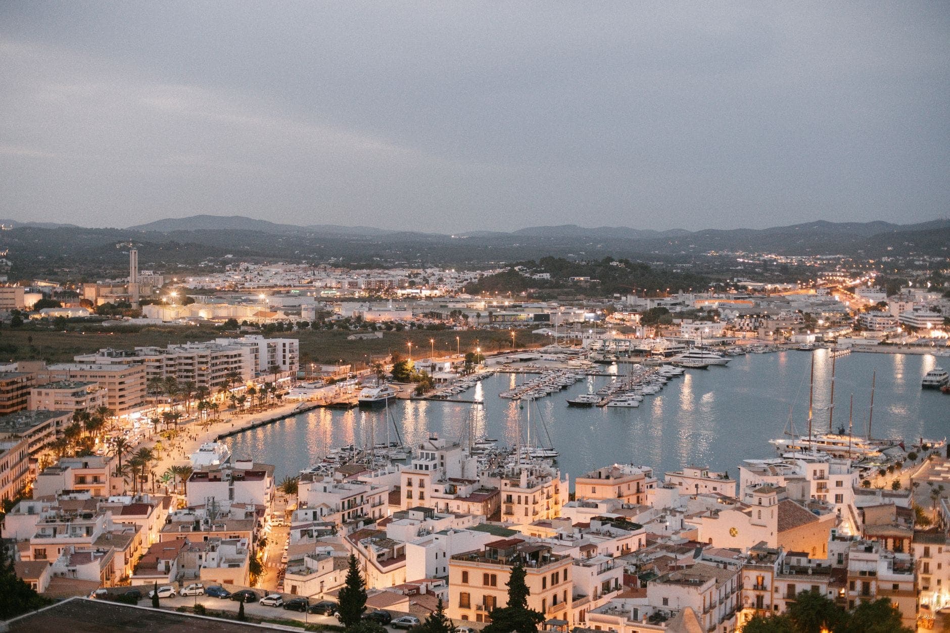 Evening view over Ibiza town showing a mix of whitewashed buildings, marina with yachts, and the wider urban landscape.