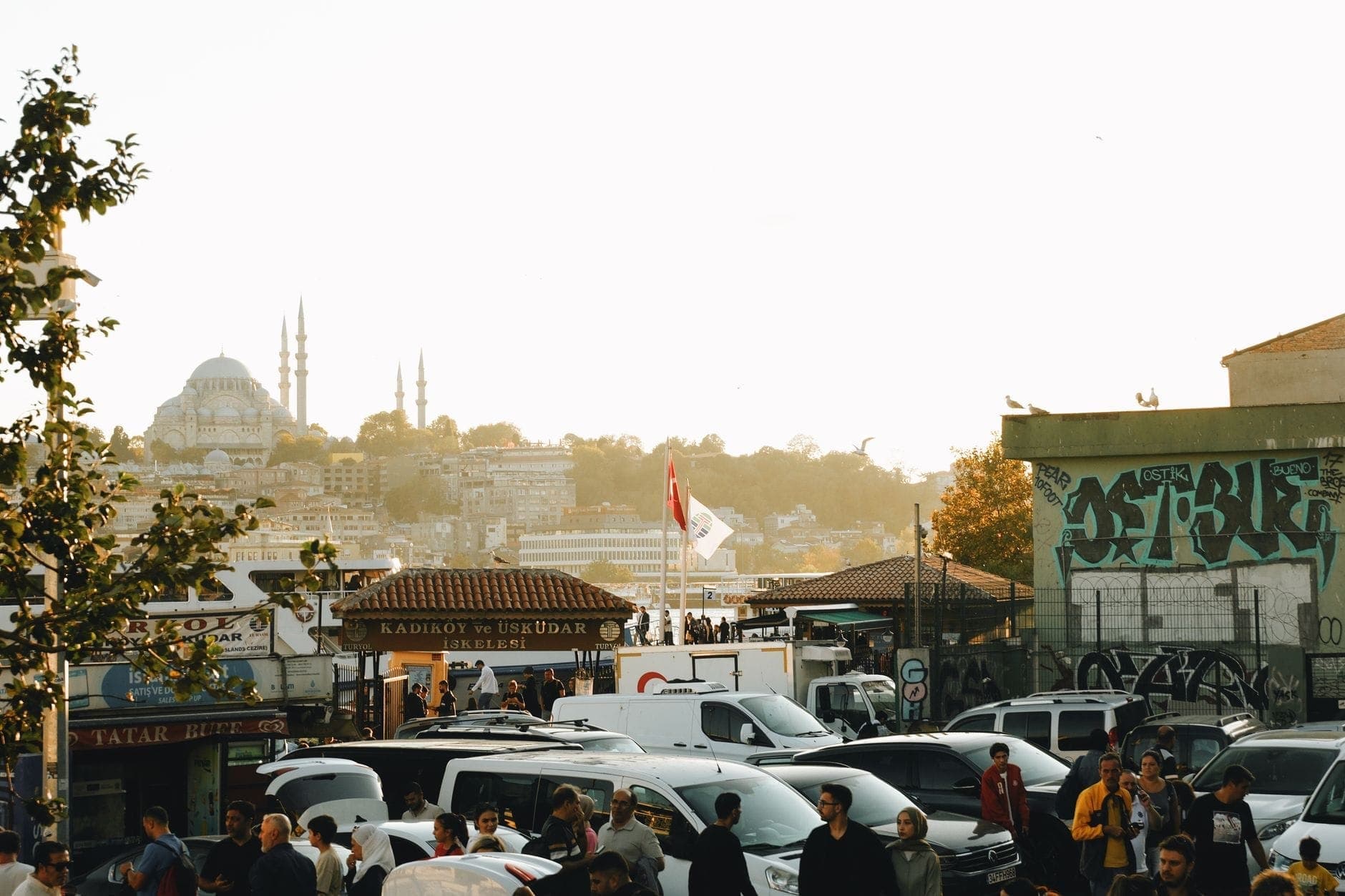 Vivace scena di strada sul lato asiatico di Istanbul con folla, automobili, bancarelle del mercato, cupole di moschee e un cartello del traghetto per Kadıköy e Üsküdar.