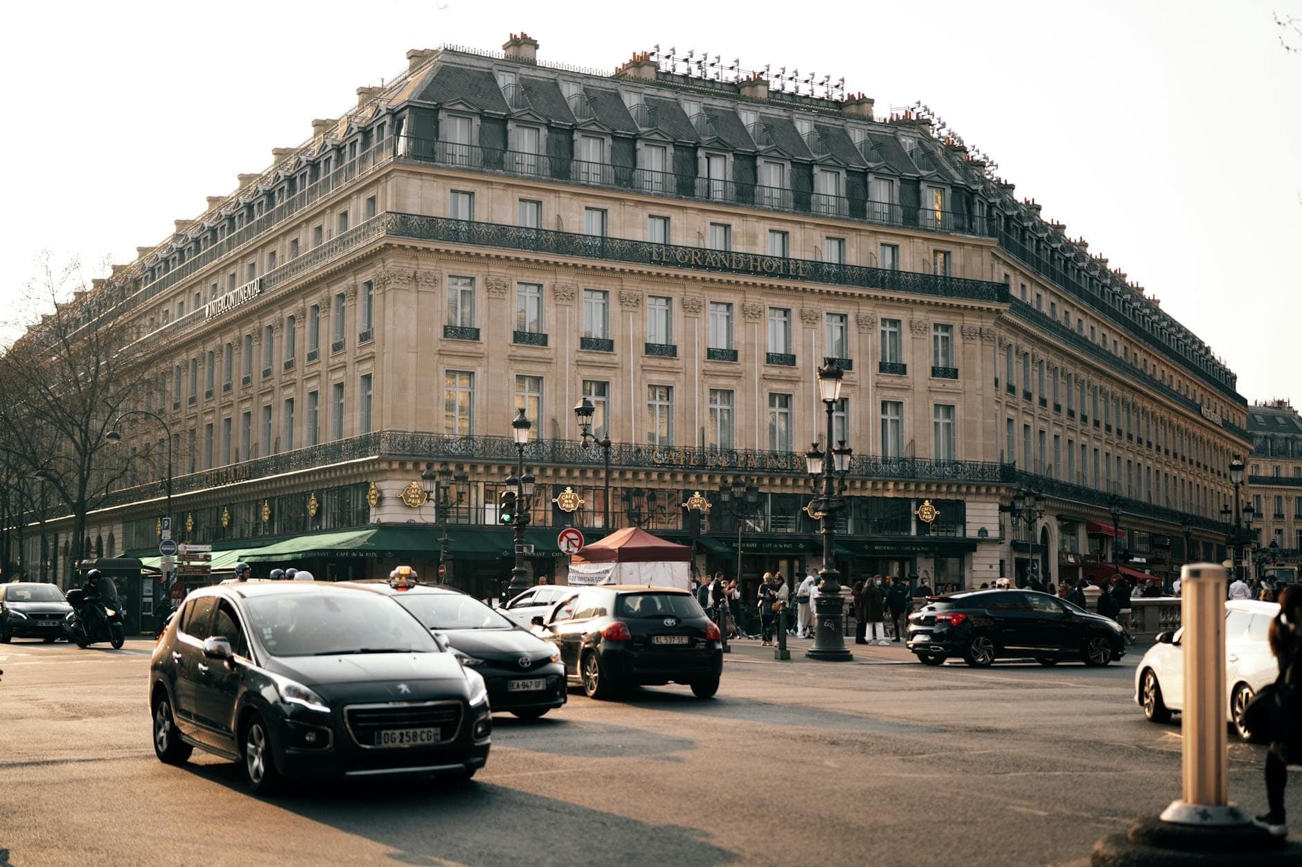 Phố Paris rộng rãi với những tòa nhà Haussmann cổ điển, xe cộ tấp nập, người đi bộ và những cây đèn đường thanh lịch trong một ngày nắng đẹp, gợi lên không khí sôi động của các khu phố Paris.