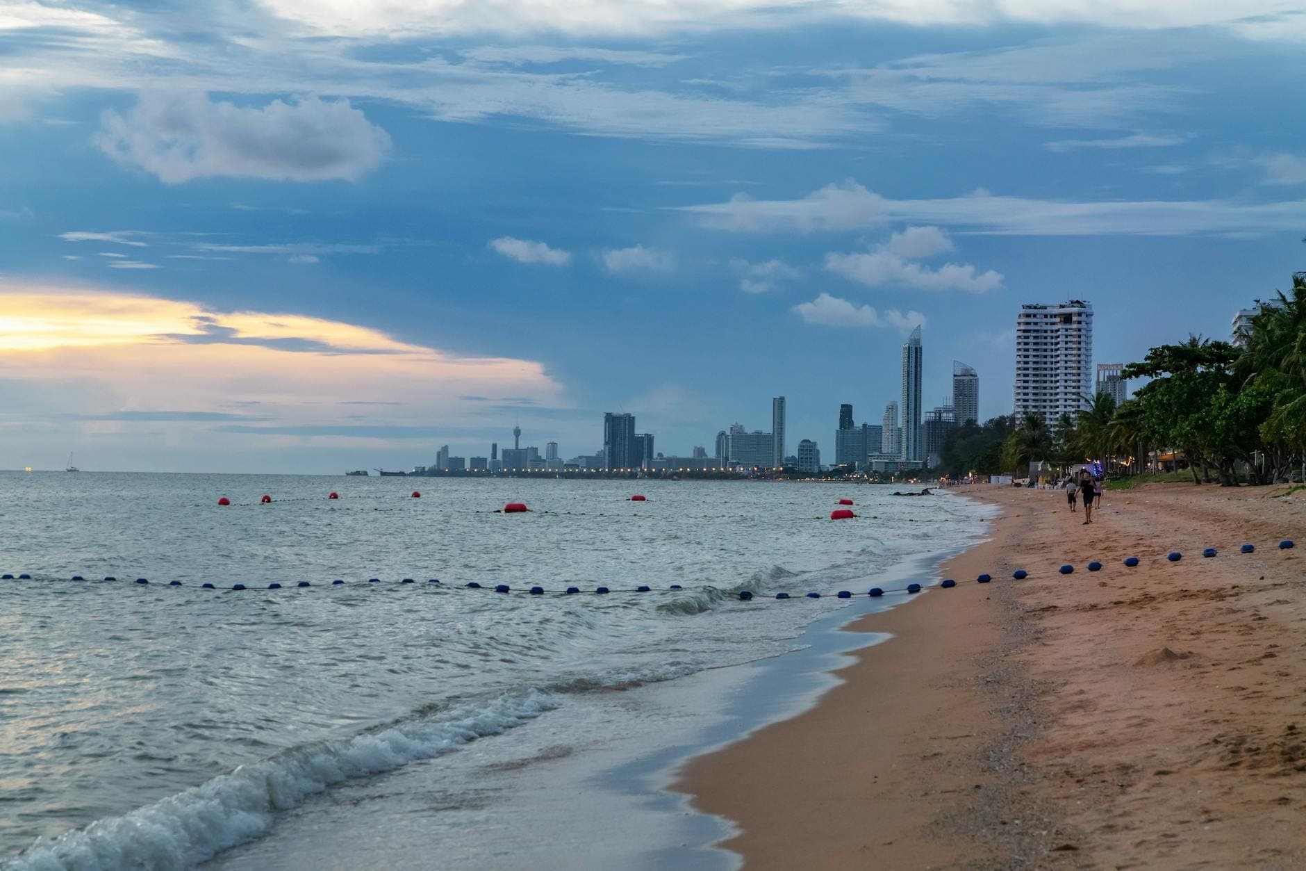 Wide sandy beach in Pattaya at sunset with calm waves, high-rise hotels in the distance, and only a few people walking along the shoreline.