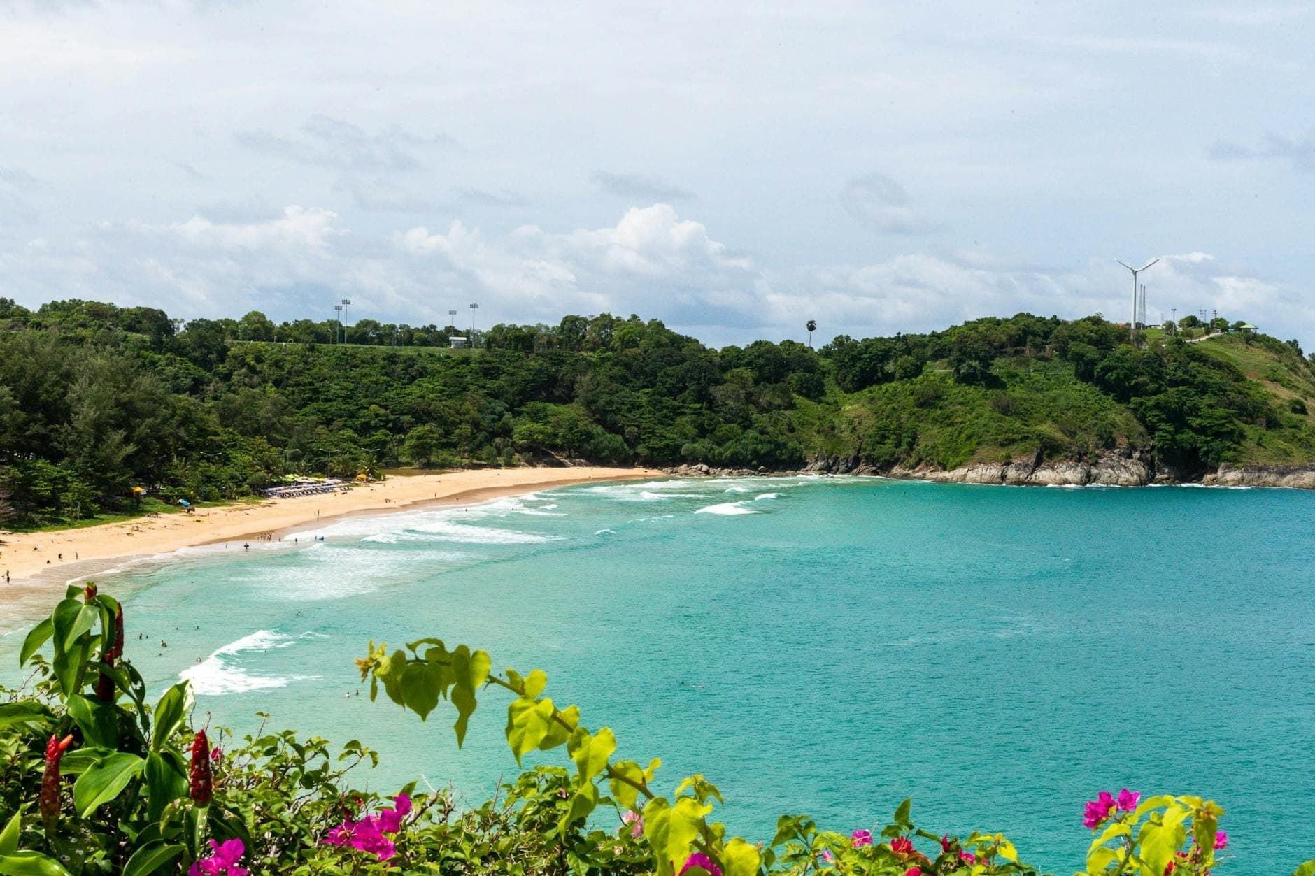 Bãi biển nhiệt đới Phuket với làn nước xanh ngọc, bãi cát vàng, đồi núi xanh tươi và những bông hoa rực rỡ dưới bầu trời có mây.