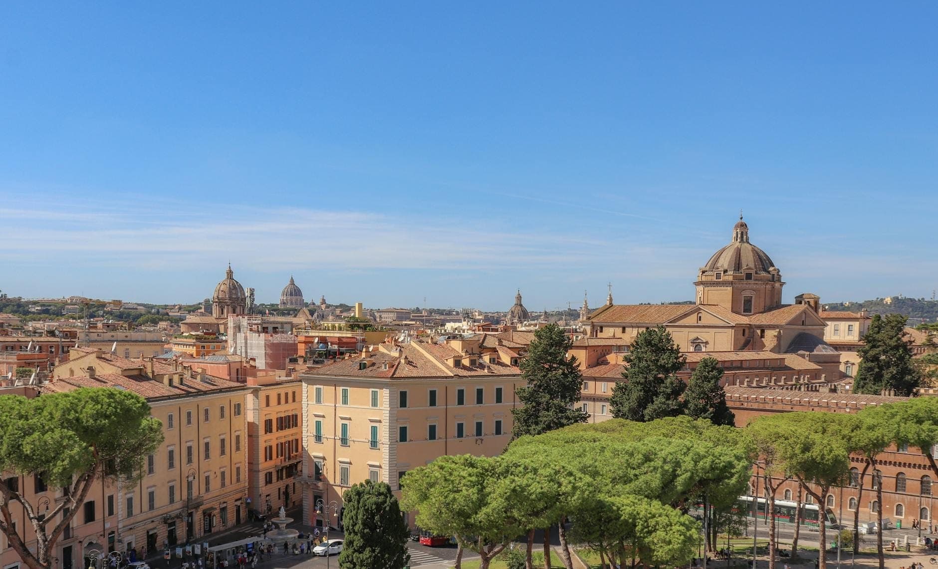 Toàn cảnh thành phố Rome với những nhà thờ vòm cổ kính, tòa nhà kiến trúc La Mã và cây xanh dưới bầu trời xanh trong, thể hiện nhiều khu phố khác nhau.
