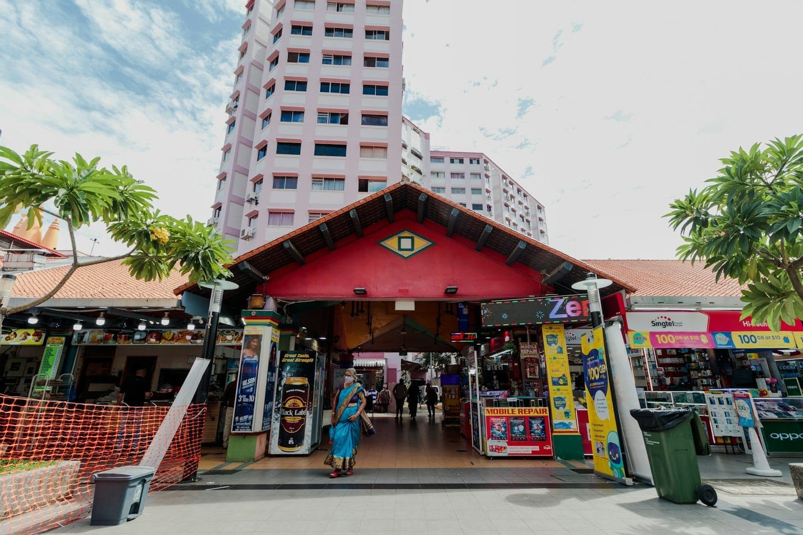 An open-air community market entrance with shops, high-rise HDB flats, and people walking, showing Singapore neighbourhood life.