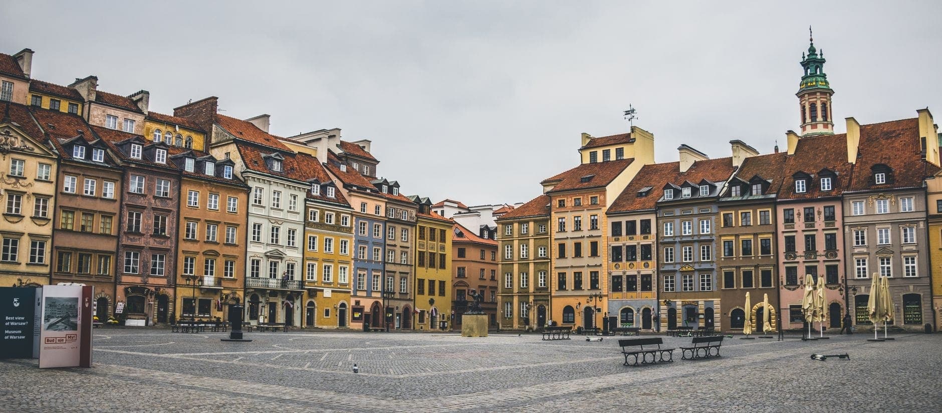 Panoramablick auf den Marktplatz der Warschauer Altstadt mit bunten historischen Gebäuden, Café-Terrassen und einem Uhrenturm an einem bewölkten Tag.