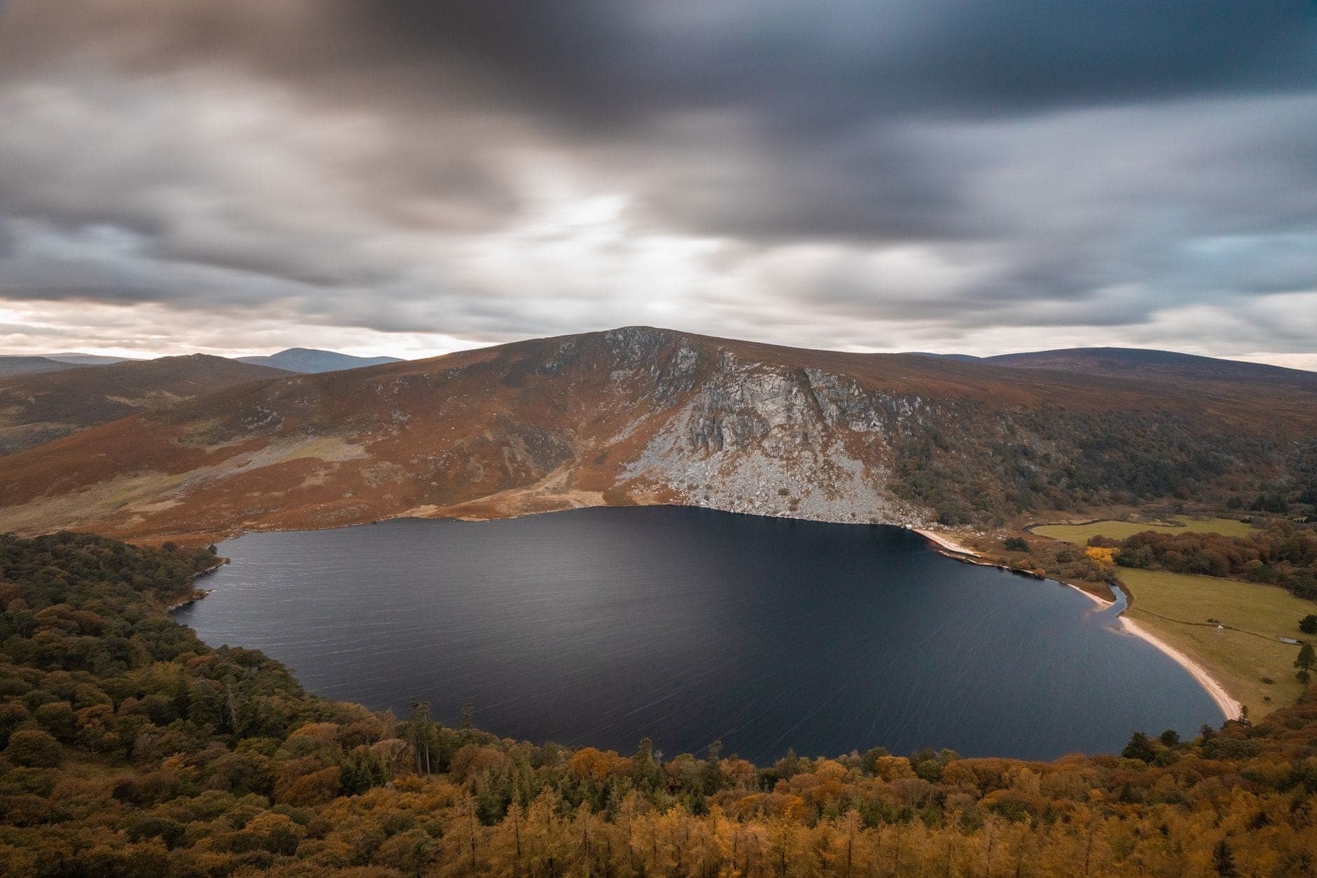 Vue grand angle spectaculaire de Lough Tay avec ses eaux sombres, ses rives boisées et ses collines escarpées dans le Parc national des Wicklow Mountains sous un ciel nuageux.