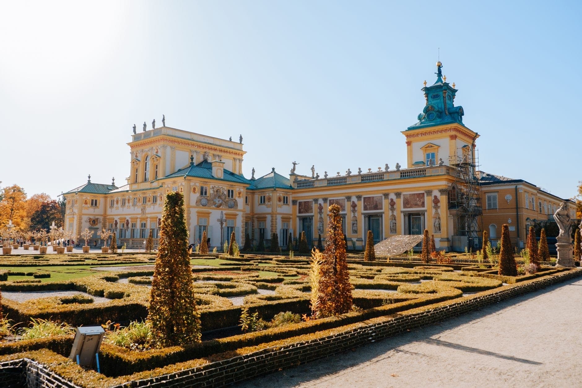 Weitwinkelaufnahme von Schloss Wilanów mit prachtvollen Barockgärten im Vordergrund; goldenes Herbstlicht und blauer Himmel betonen die gelbe Fassade und das grüne Dach des Schlosses.