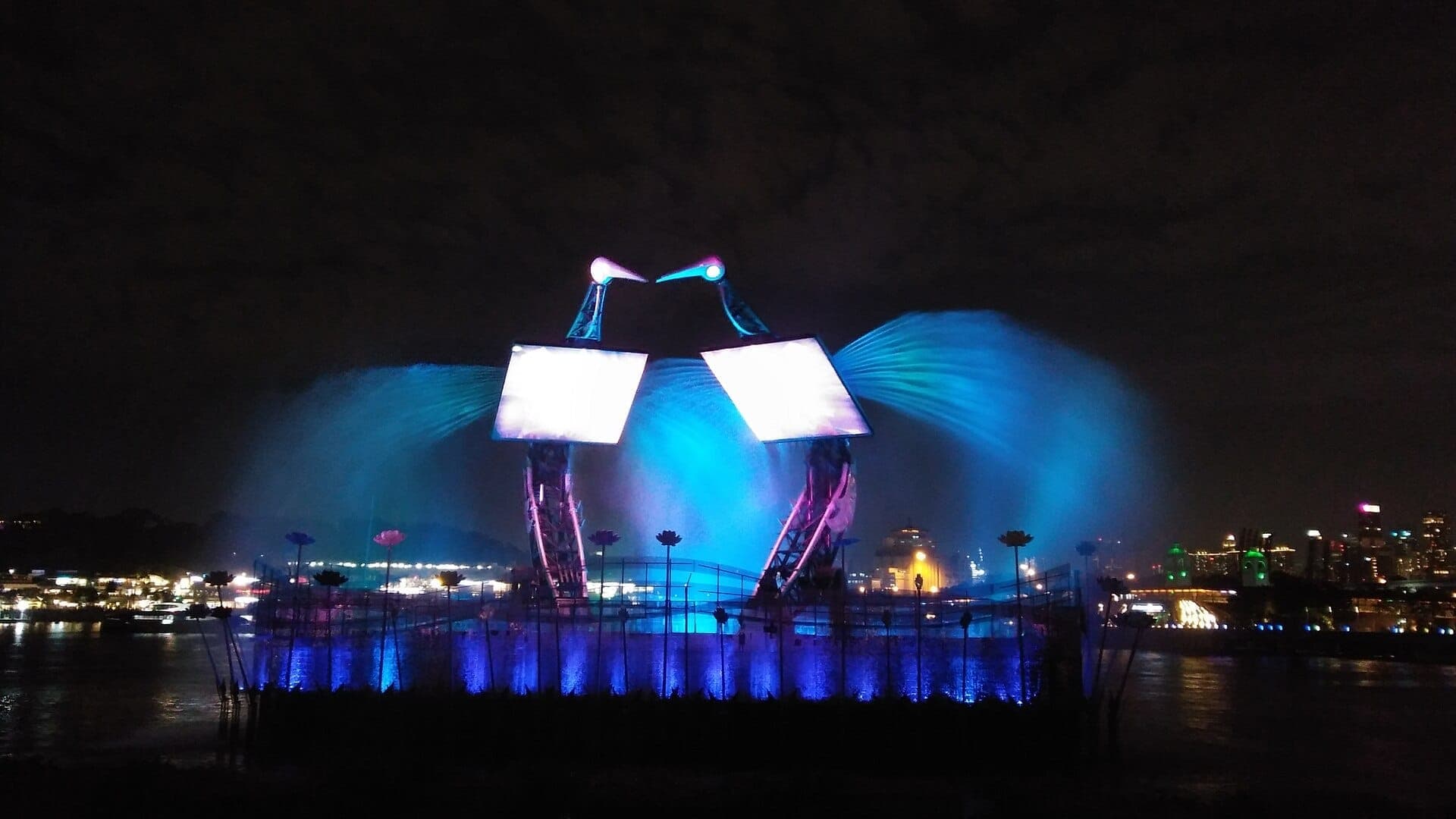 Colorful lights and water jets illuminate the Wings of Time outdoor stage at Sentosa, with a nighttime cityscape in the background.