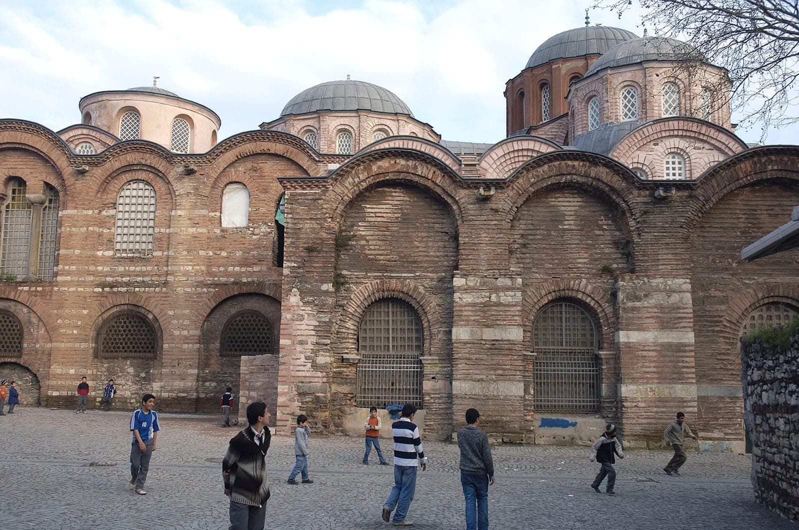 Außenansicht der Backsteinmauern und Kuppeln der Zeyrek-Moschee, mit Kindern, die an einem hellen Tag im gepflasterten Innenhof spielen.